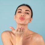 Close beauty portrait of topless woman with perfect skin and natural make-up, plump nude lips, on a blue background, sends a kiss